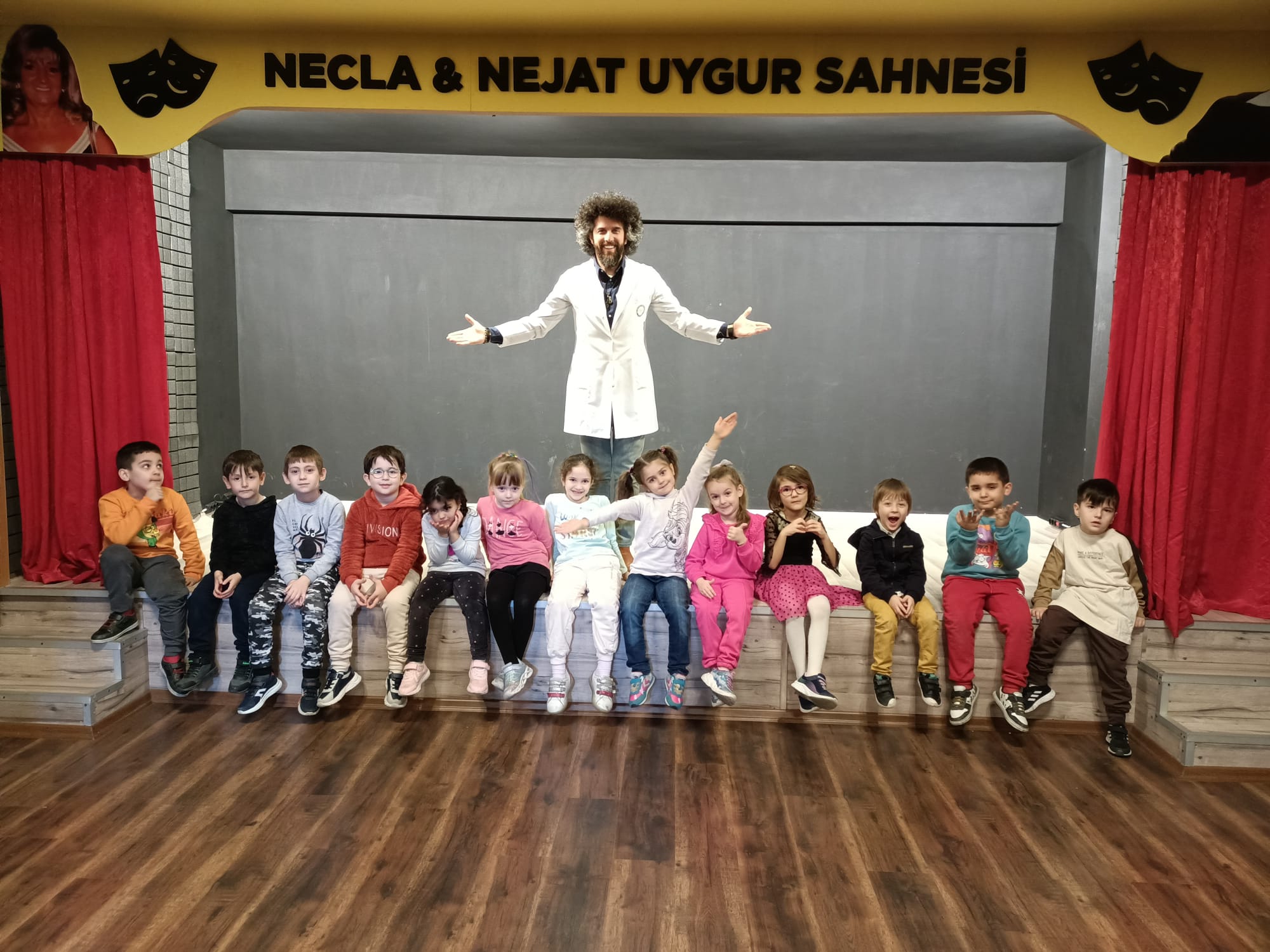 İlyas Çağlayan sahnenin ortasında, kollarını açmış; önünde öğrencileri oturuyor