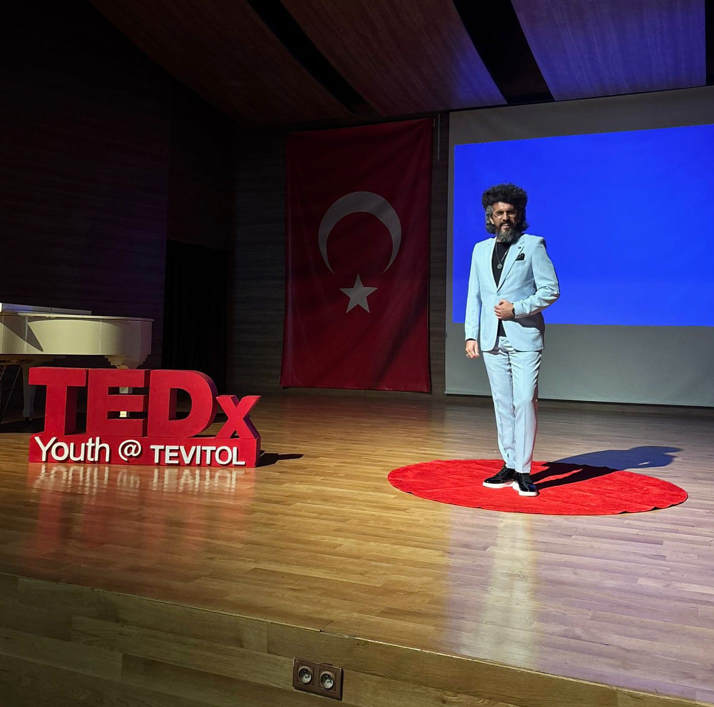 İlyas Çağlayan TEDx sahnesinde tam boy; sahnenin geniş açısı ve TEDx harfleri birlikte görünüyor